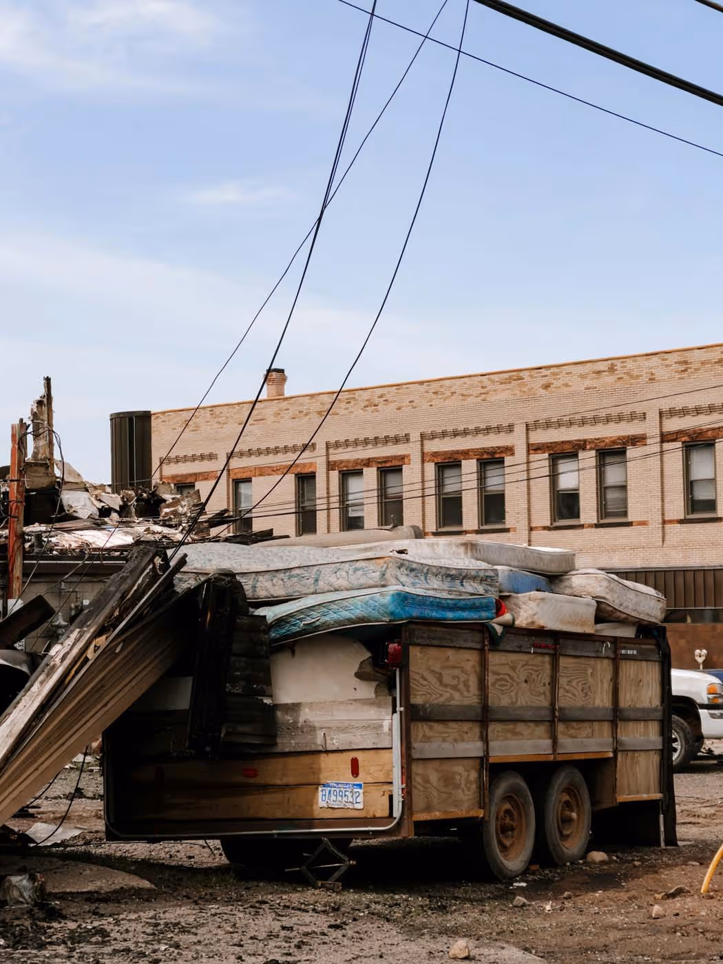 Truck trailer loaded with old mattresses and debris parked in front of a damaged building and another beige brick building.